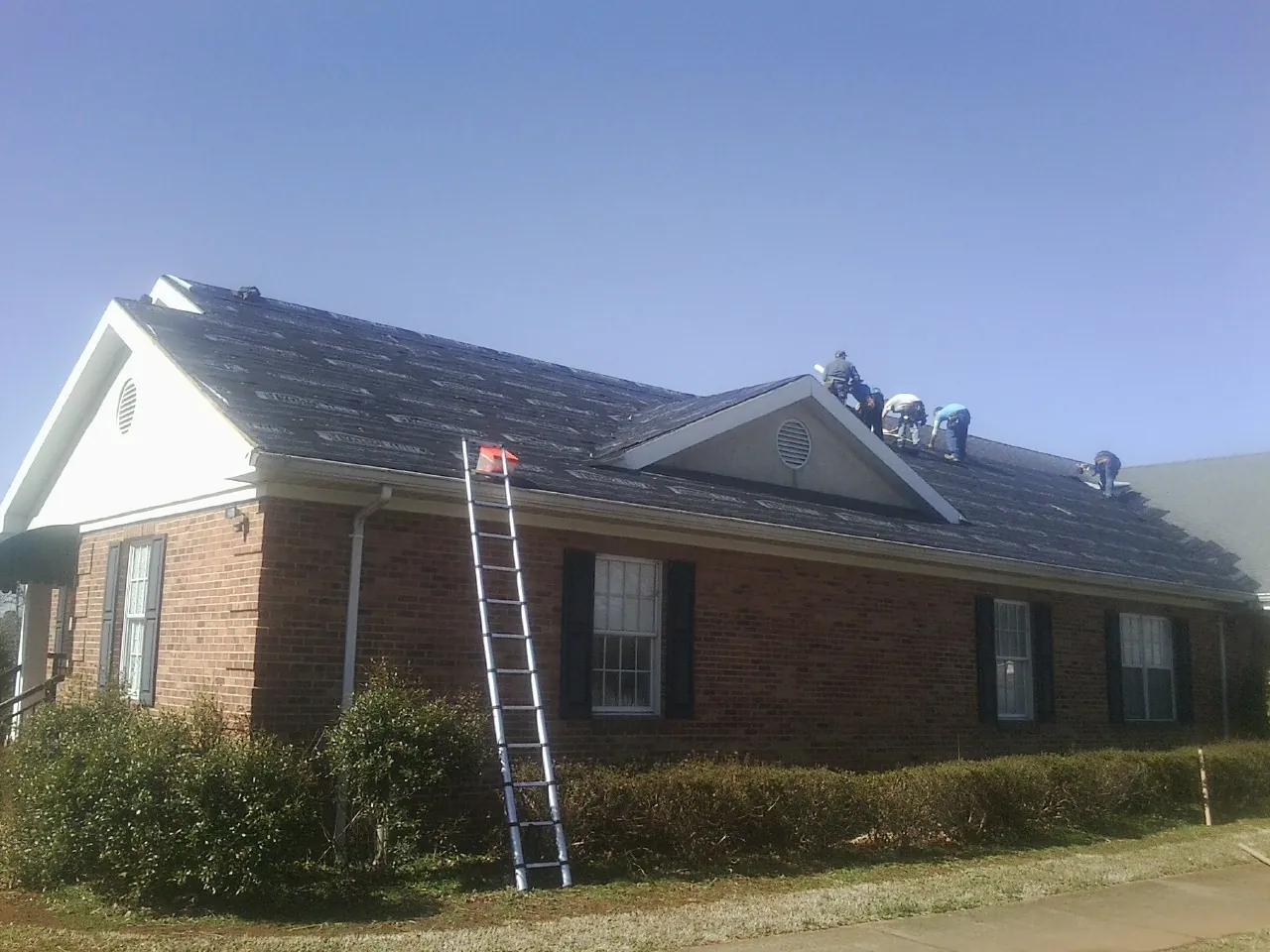Crew tearing shingles off a brick ranch roof with a ladder set — Metal Roof Repair work by our crew in Wysox