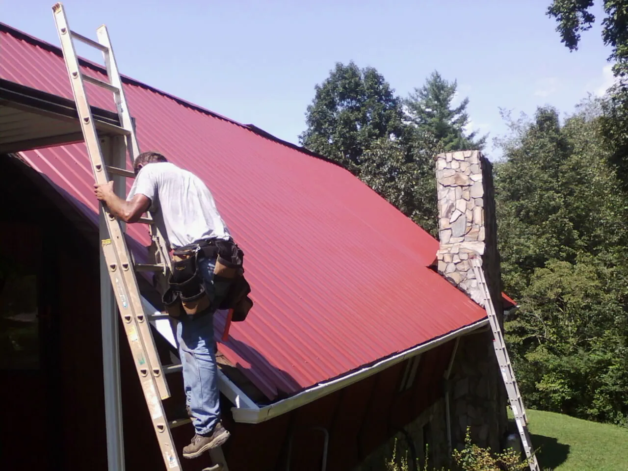 Roofer on a ladder beside a red metal roof and stone chimney — Soffit & Fascia Repair by Roofing Brothers in Wysox